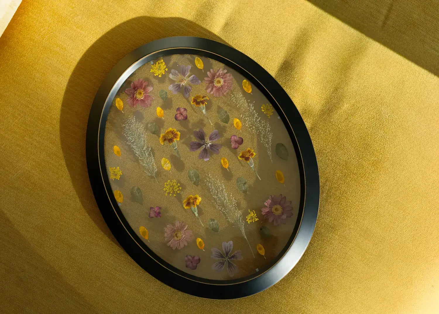 Oval wood frame with unique arrangement of pressed flowers on a yellow fabric background