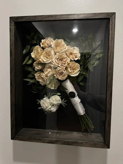 Preserved wedding bouquet and matching lapel flowers in a dark wooden frame against a white wall